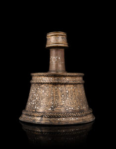 M3200-0217_Chandelier mamelouk en laiton incrusté d'argent au nom d'un grand émir (officier) du Sultan al-Nasir Muhammad ibn Qalawun_Egypte, 1293-1341_De forme typique avec une base tronconique, un col tubulaire et une bobèche cylindrique inclinée, une épaule concave, à décor incisé et incrusté d'argent et de pâte noire. Le corps est décoré d'une inscription monumentale en thuluth divisée par deux médaillons floraux, le tout sur un fond de rinceaux ondulants et spiralés, et est encadré de bandeaux mineurs à décor de chevrons en relief et de feuilles stylisées séparées par une bande de rinceaux ondulants. L'épaule est avec une inscription radiale en thuluth ponctuée de deux feuilles de lotus épanouies, et cernée du même registre de feuilles stylisées séparées par une bande de rinceaux ondulants. La bobèche est inscrite d'une phrase en coufique ponctuée de trois rosettes à six pétales. Le col reprend ce motif et celui de la fleur de lotus épanouie associé à des motifs flammés. Base de la panse avec une fêlure, bon restes de l'incrustation d'argent. H. 27 cm