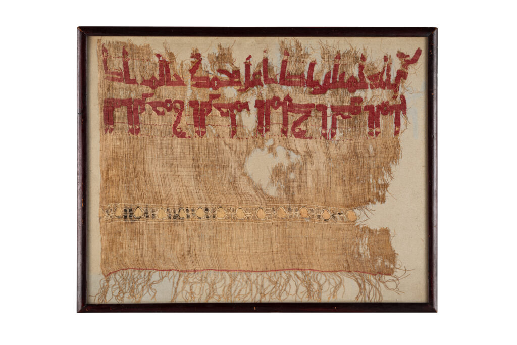 M3200-0240_Adjugé 14 000_Grand fragment de tiraz fatimide_Egypte, IXe siècle_En lin uni non teint, tissé avec une bande de lin rouge de deux grandes inscriptions en coufique, disposées en symétrie, avec d'élégantes verticales étendues contenant des carrés blancs. En dessous, une ligne de cercles accompagnés de lignes spiralées, en blanc et noir. Sur la partie inférieure, ligne rouge et franges. Cadré sous verre. 33 x 44 cm sans les franges. Provenance : Collection Jean-Charles Tauzin (1889-1957)