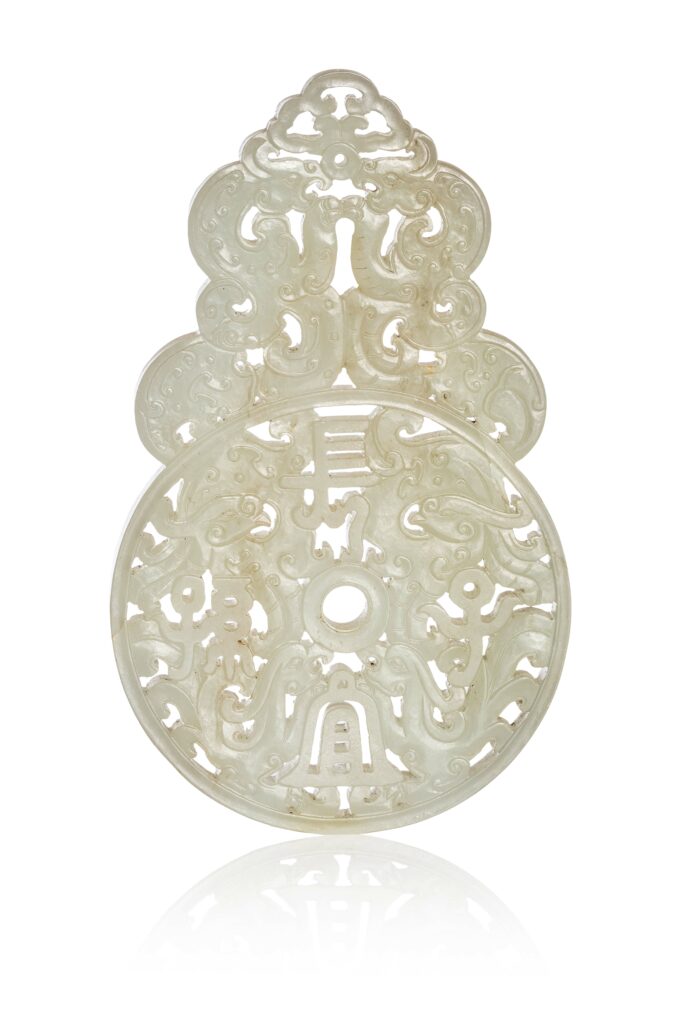 N0456-0012_CHINE, XVIIIe siècle Élément en jade céladon À décor sculpté et ajouré prenant la forme d'un disque bi comprenant quatre phénix stylisés autour d'un caractère, la partie supérieure ornée de dragons stylisés. La pierre aux tons céladon légèrement infusés de rouille. Hauteur : 12,7 cm Largeur : 7,7 cm Adjugé 28 000 €