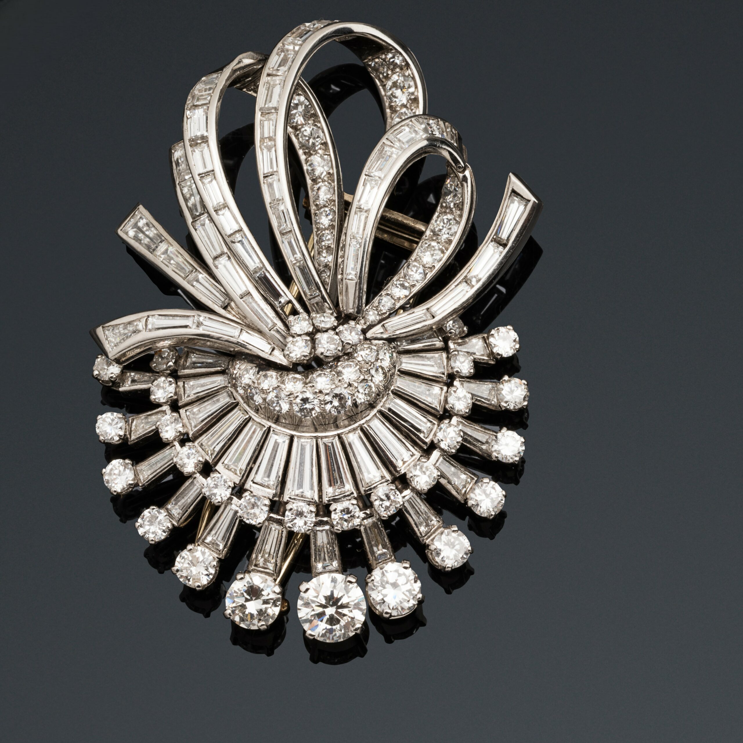 Broche diamants, Clip de revers en platine (950 millième) et or gris 18k (750 millièmes) figurant un motif rayonnant retenant des rubans noués, l'ensemble serti de diamants ronds taille brillant, baguette et tapers. Travail français vers 1950. Poids brut : 39,6 g. Haut.: 6,2 cm Adjugé 8 000 €